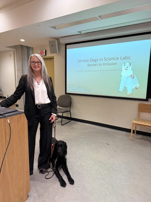Joey is a female with long gray hair standing casually next to a podium wearing a black business suit and white bouse and glasses. She is smiling. Pax is lying next to her wearing a red service dog vest. Behind them is a screen with a drawn photo of Sampsons wearing a white lab coat and safety goggles, and boots. The presentation is titled, "Service Dogs in Science Labs: Barriers to Inclusion." 