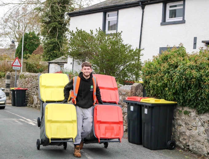 A man in an orange high vis jacket pulls four wheeled recycling bins with yellow or red lids along a road in front of houses with  stone walls.