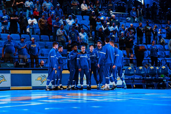 A group of athletes in matching blue tracksuits stands in a circle on the mat at South Dakota State University, surrounded by cheering spectators. The court features a jackrabbit logo, and the scene captures a moment of team unity and anticipation before the wrestling match 