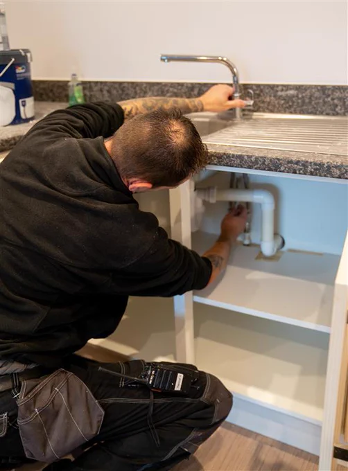 A man wearing a black fleece and work trousers fixing a pipe under a sink.