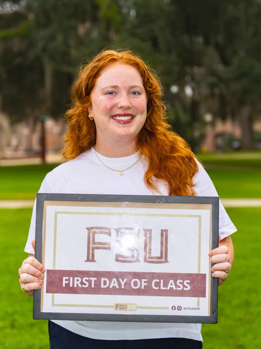A student holding a sign displaying "FSU First Day of Class" while standing on a grassy area with trees in the background. The person has red hair and is smiling.