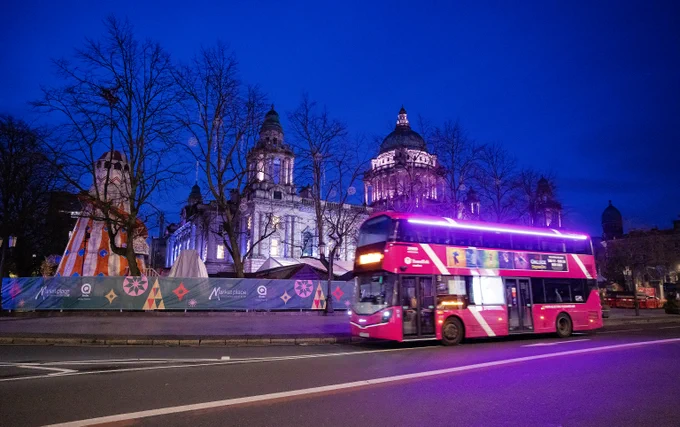 Photo shows a pink metro bus passing the City Hall and Christmas market at night time. 