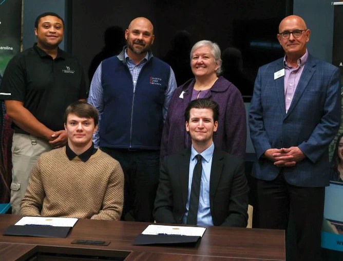 Group of six individuals posing for a photo at a professional event, with some participants standing and others sitting at a table, all smiling.