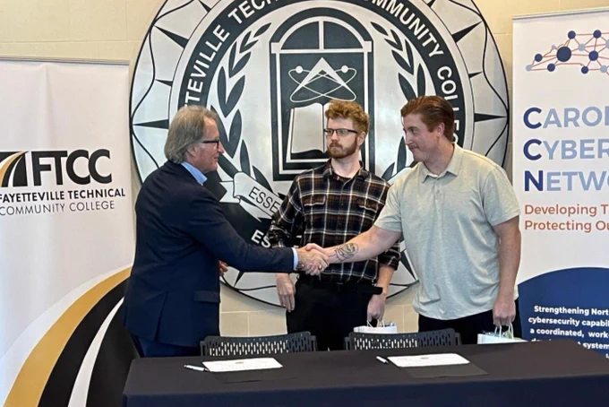 Three individuals participate in a handshake at Fayetteville Technical Community College, standing in front of banners with the college's logo and the Carolina Cyber Network logo.