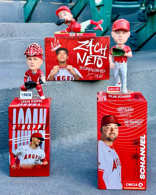 Three individual bobbleheads depicting Logan O'Hoppe, Zach Neto, and Nolan Schanuel are displayed on the stairs next to the stands at Angel Stadium. The three bobbleheads sit atop their boxes, showcasing the name of the Angels player on the red designs. 