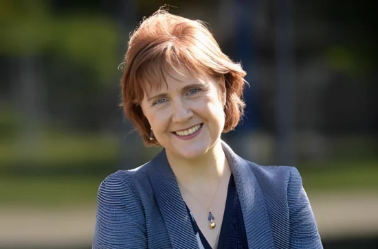 A woman with short, auburn hair smiles warmly at the camera. She is wearing a navy blazer over a dark top and a pendant necklace. The background is blurred greenery.
