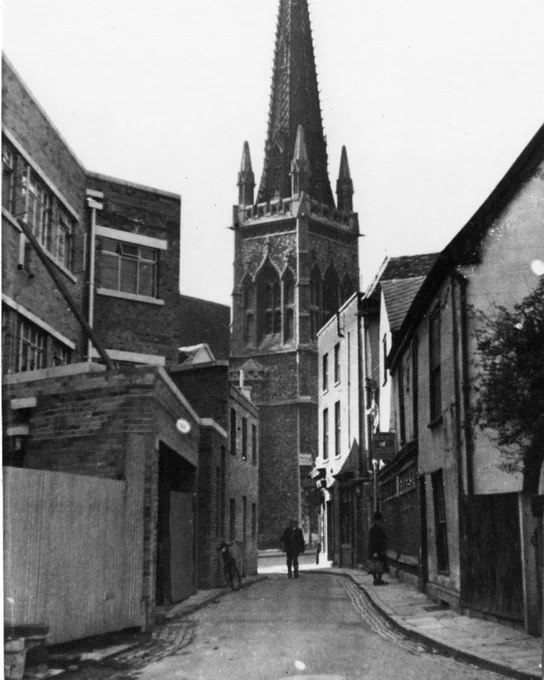 Black and white photograph of a narrow street lined with two and three storey buildings, with a tall Gothic church tower and spire filling the background. A bicycle leans against a brick building on the left and two figures walk along the street towards the camera.