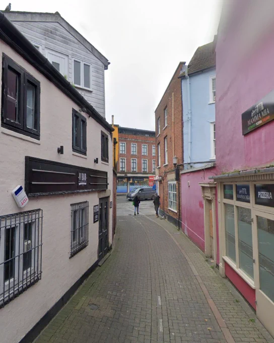 Colour photograph taken from the same narrow street, showing a gently curving paved lane between closely packed buildings. The shopfronts include a bar and a pizzeria, painted cream, pink and blue, with barred windows and modern signs. In the distance a small square opens out with taller brick buildings and parked cars, and two people walk away from the camera.