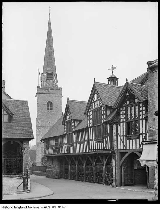 A black and white photograph of a timber framed building with gables on the right, and the tower and spire of a church on the left. To the left again is part of a building with a tiled roof. The street is empty, save for a parked bicycle leaning against the front of the timber framed building.
