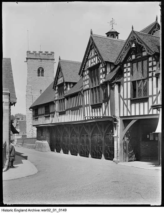 A black and white photograph of a timber framed building with gables on the right, and the tower of a church to the left. A woman stands on the pavement on the street corner on the left of the photograph.