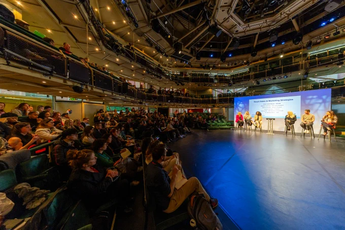 Large audience attentively watching a panel discussion on stage in a modern auditorium with multiple seating levels.