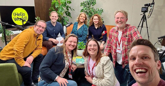 Group of seven people pictured in a podcast studio. There are potted plants and a Hello World sign in the background.
