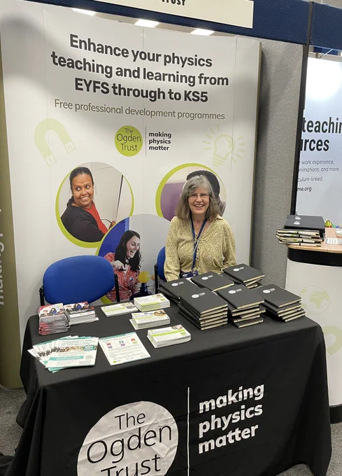 The Ogden stand at ASE a display saying Enhance your physics teaching and learning from EYFS through to KS5 with a table in  front covered with a black table cloth featuring the Ogden logo and leaflets and notebooks on the table. A lady is sat behind the table ready to talk to visitors to the stand! 
