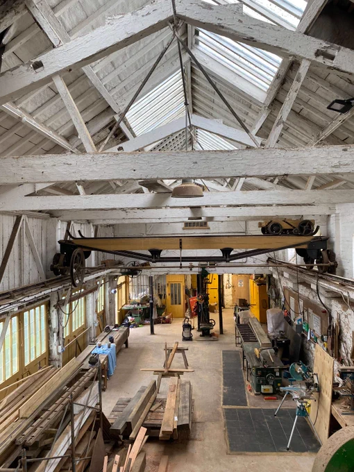 A view of a historic joiners shop from above. The ceiling has wooden white beams across it with glass windows letting light in. Below are large pieces of wood and machinery. 