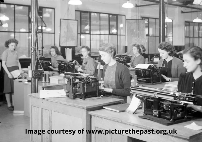 An office with rows of wooden desks each with a large typewriter sitting on it. Each desk has a female secretary sitting at it typing. They are all smartly dressed in neat dresses or blouses and cardigans.