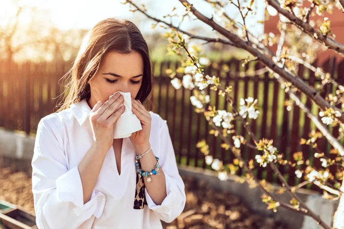 Woman using a tissue outside near blooming trees, experiencing allergies.