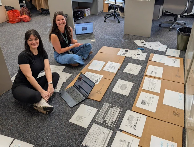Two women are seated on a floor with one holding up a laptop computer. There are printed pages of information sprawled out in front of them.
