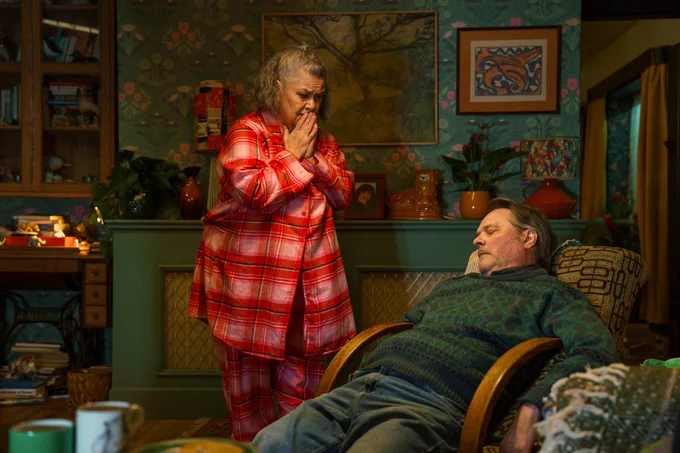 Dawn French's character, dressed in pyjamas, looks distressed while standing over Mark Heap's character, who is slumped in a chair unconscious