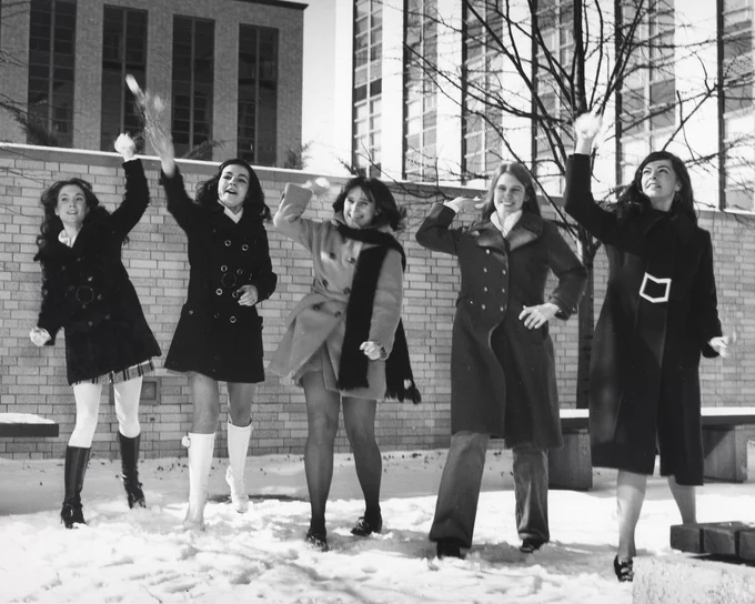 Black and white image of five smiling women wearing winter coats and throwing snowballs. They are standing outside in the snow with a brick wall and buildings behind them.