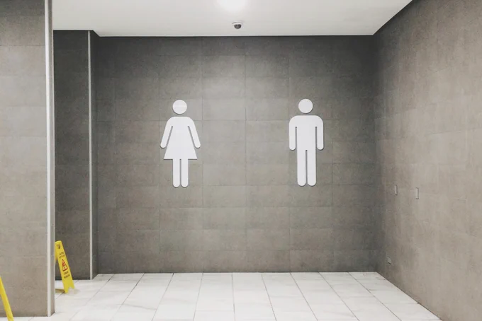 Wall with male and female restroom symbols on a gray tiled background. Floor is white, with a yellow caution sign in the corner.