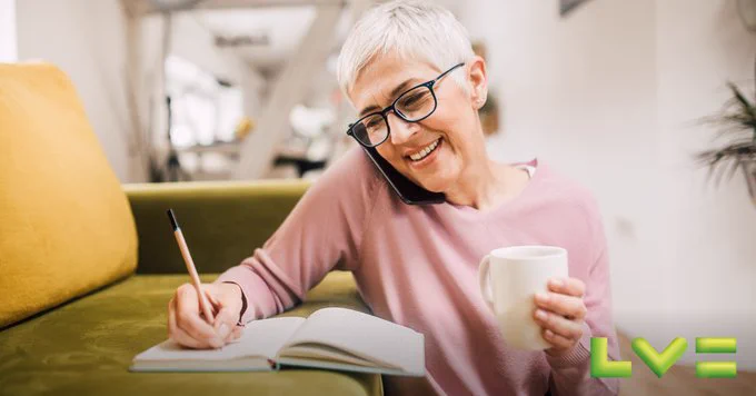 A smiling lady with glasses and a pink jumper is on a phone call. She is writing in a notebook and holding a mug, leaning against a green sofa. The LV logo is a the bottom of the image.