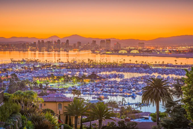 Sunset view of San Diego skyline and marina filled with boats, with vibrant orange and purple hues in the sky.