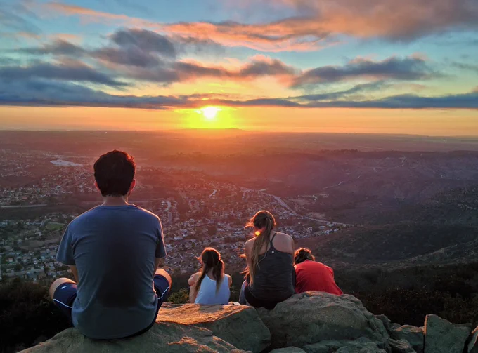 Four people sitting on a rock overlooking a scenic sunset above a vast landscape.