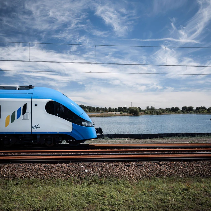 An "elf" train running along tracks near a large body of water with a clear sky above.