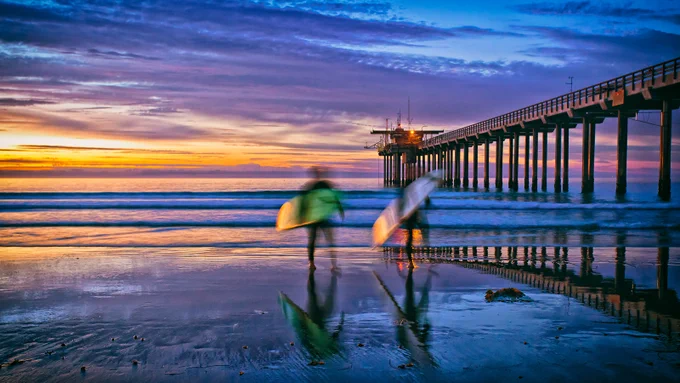 Two surfers walking along the shore near Scripps Pier at sunset, with vibrant sky reflections on the wet sand.