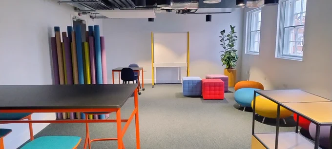 The media / maker space room at Tavistock Place. The room features colourful furnishings, along with a moveable whiteboard and funky shelving. A plant can also be seen in the corner.