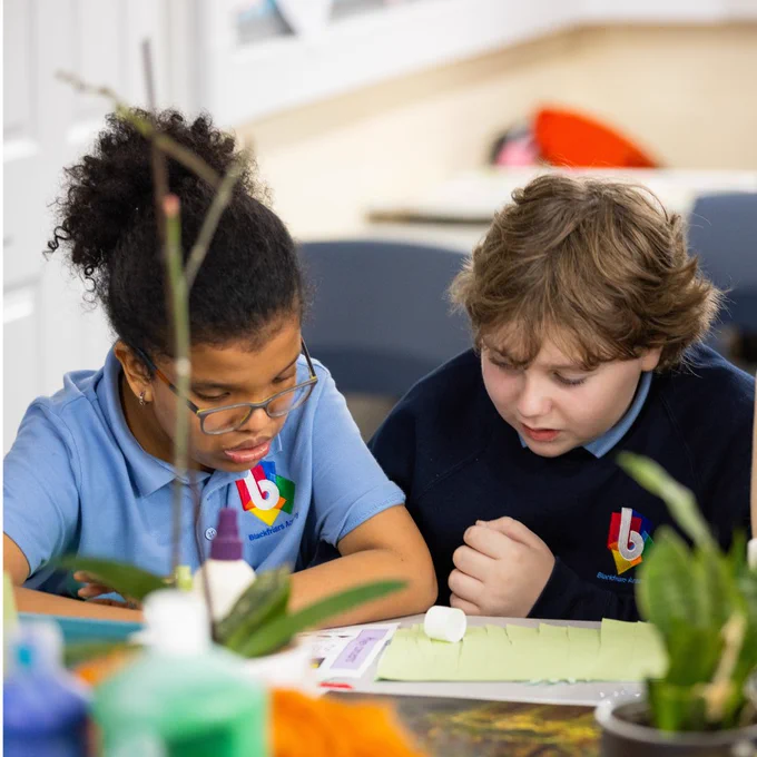 Two children in blue school uniforms, aged around 12 or 13-years old, sit in a classroom. They look down at a worksheet on the table in front of them.