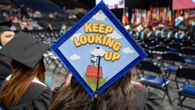 A graduate wears a blue-bordered mortarboard featuring Snoopy on his doghouse with the phrase “Keep Looking Up,” with empty chairs and flags visible in the background of the graduation venue.