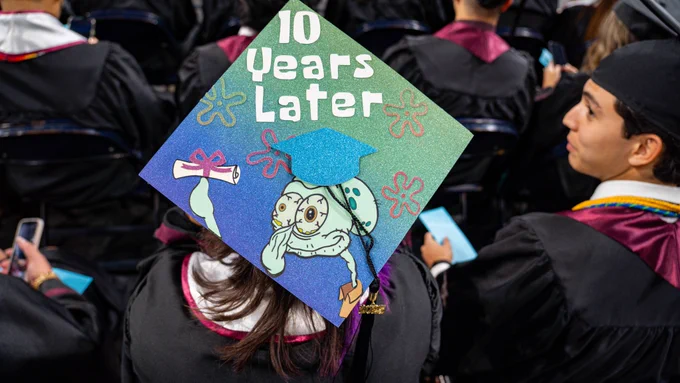 A graduate wears a colorful mortarboard decorated with a cartoon character Squidward and the phrase “10 Years Later,” sitting among rows of graduates during a commencement ceremony.