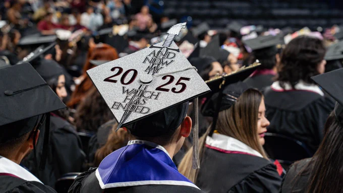 A graduate in a black gown wears a silver glitter mortarboard reading “And now my watch has ended” with the year “2025” and a sword graphic, seated among fellow graduates at a commencement ceremony.