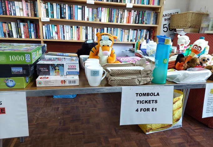 image of the tombola table and sign "Tickets 4 for £1" with a backdrop of book shelves. 