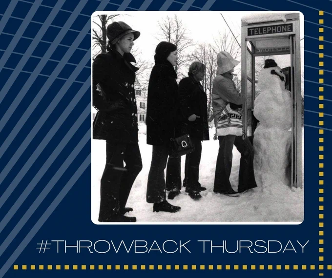 A black and white photograph of students waiting in line outside a phone booth occupied by a snowman.