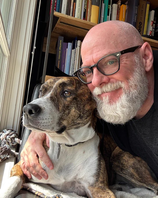 Dan Rhatigan smiling while holding his dog, both sitting next to a window. Bookshelves are in the background.