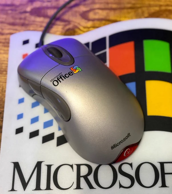 Image is of an older, wired Microsoft Office mouse resting on a mousepad with a vintage Microsoft logo on it.