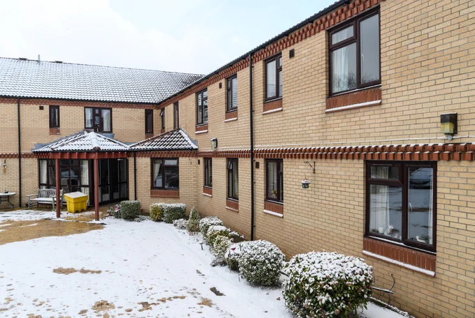 A photo of the exterior one of our Housing schemes with a dusting of snow. It is a two-storey brick L-shaped building with a courtyard in front of the entrance.