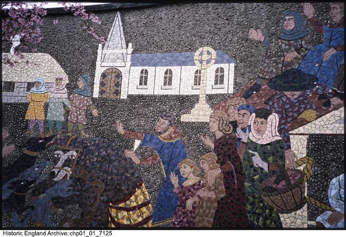 A colour photograph of a mosaic depicting a market scene. In the foreground is a group of people, some with baskets and hats, watching a group of dogs attack a bear. In the centre of the image is a market cross, and beyond this is a church. To the left, three people stand in front of another building.