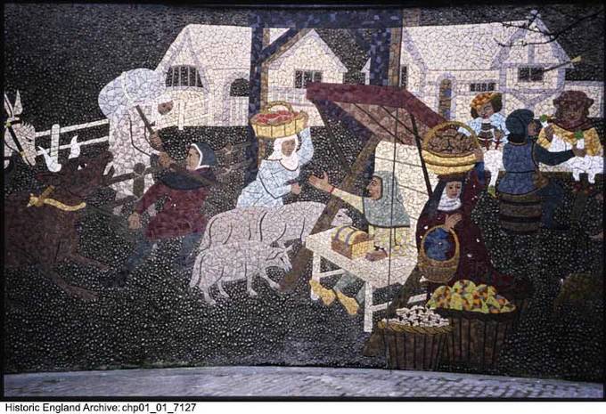 A colour photograph of a mosaic depicting a market scene. In the centre is a woman holding a basket of fruit on her head, with three sheep at her feet. To the right is a person seated in a stall with a low table in front of them. This scene is surrounded by people with farm animals, baskets and produce. In the background are two buildings of pale stone.