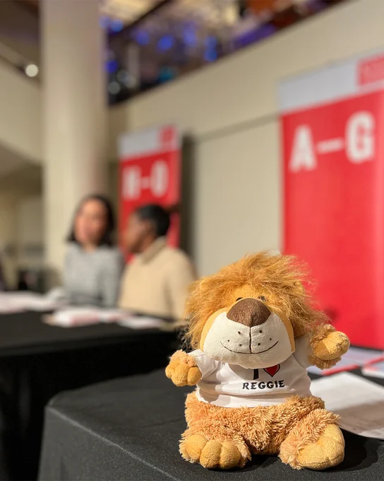 A cuddly toy version of the King's mascot, Reggie the Lion, sits on a desk. In the background are the registration desks for graduations at the Royal Festival Hall.