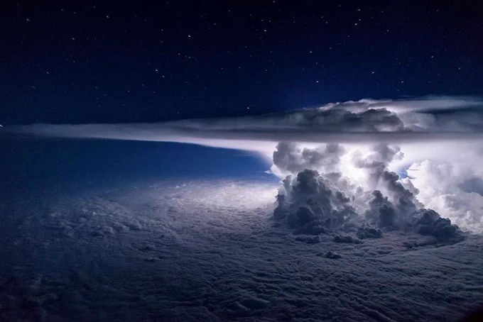 A massive cumulonimbus storm cloud rises from the lower cloud deck into the stratosphere. The image captures a lightning flash within the storm, illuminating the massive cloud structure, including its turbulent updraft and smooth, flat "anvil" top. 