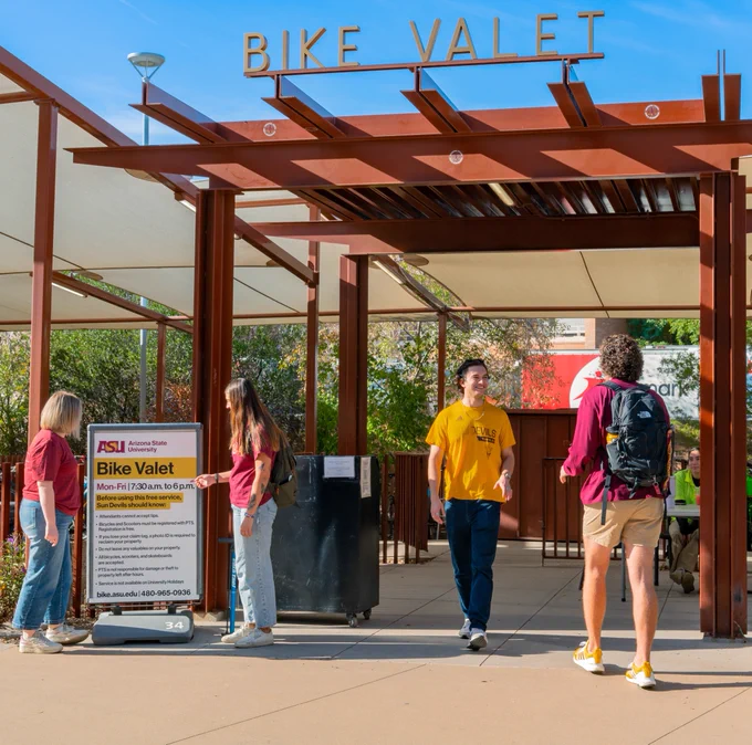 Outdoor bike valet station with a metal canopy and four people interacting near a sign detailing services at Arizona State University.