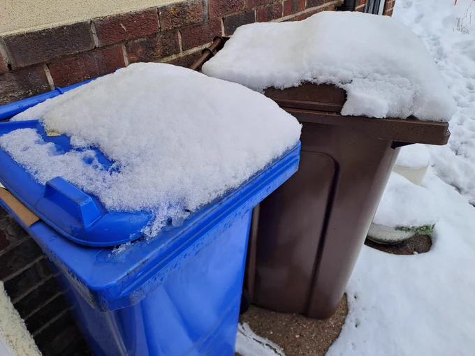 Snow topped blue bin and brown bin standing on snowy ground next to a house.