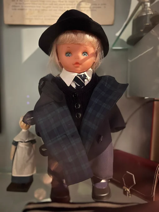 A close up of a museum doll on display at Hollytrees Museum, with painted eyes, pale skin and neatly styled hair, standing upright in a case and staring slightly past the camera.