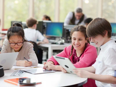 Three students collaborating at a round table using a tablet and notebooks in a bright classroom setting.
