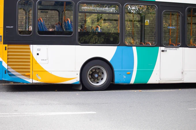 The back half of a bus (from windows down), including the back wheel with a road in front. The bus is white with teal, light blue and mustard yellow decorations. 
