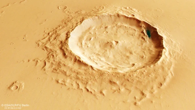 A close-up view of a large impact crater on Mars. The crater has steep, sharply defined walls and a wide, flat floor with uneven ridges. Surrounding the crater is a gently sloping, textured surface in shades of light tan and reddish-brown. A small patch of darker material appears near the upper right inner wall of the crater.