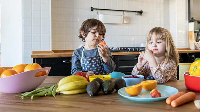 Two children are sitting at a kitchen table, eating fresh fruits and vegetables. Colorful bowls and plates with various healthy foods like bananas, avocados, carrots, and oranges are spread out in front of them.